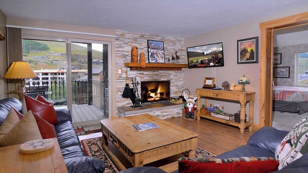 A cozy living room with a fireplace, leather sofas, a wooden coffee table, and a view through large windows. - The Bluebell, Chateaux Condo, Pool, Hot tub, Sauna (Mt. Crested Butte)