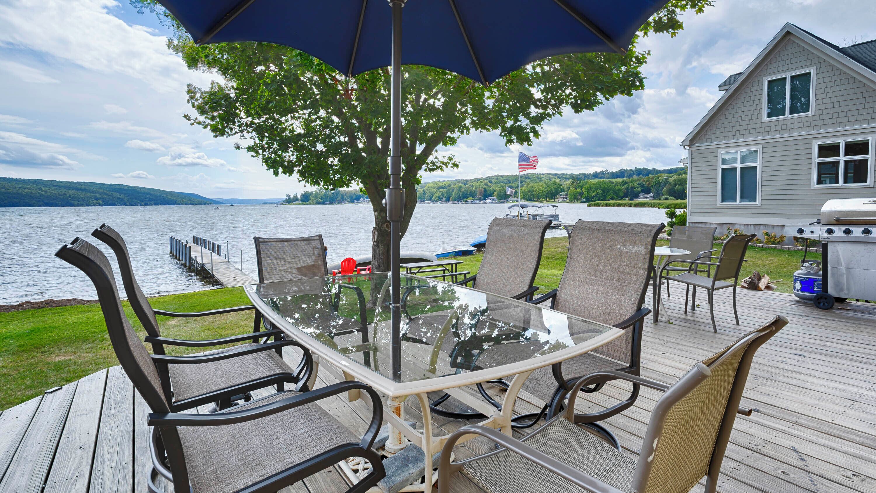 A deck with a glass table and chairs, overlooking a lake with boats and a house in the background.