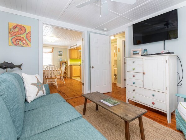 A living room with a teal sofa, a wooden coffee table, a white cabinet, and a television on the wall. - Mellow Yellow | Pet Friendly, Cottage, Fenced Yard (Tybee Island)