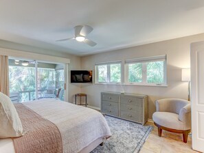 A bedroom with a bed, a chair, a TV, a ceiling fan, and a window with a view of trees.