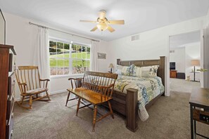 A bedroom with a bed, rocking chairs, a ceiling fan, a window with a view, and a door leading to another room.