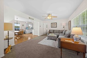 A living room with a sofa, a coffee table, a side table, a ceiling fan, and a view of the kitchen.