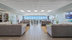 A spacious lounge area with sofas and a coffee table, a bookshelf, and a large window offering a view of the beach. - Phoenix VI 811 | Beachfront | Pickleball | Pools | My Beach Getaways (Orange Beach)