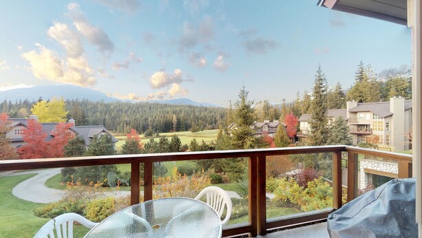 A balcony with a glass table and chairs, overlooking a residential area with houses, trees, and a mountain in the distance. - 3BR | Blackcomb Greens | Wood Burning Fireplace | Complex Hot Tub | Parking | Mountain Views (Whistler)