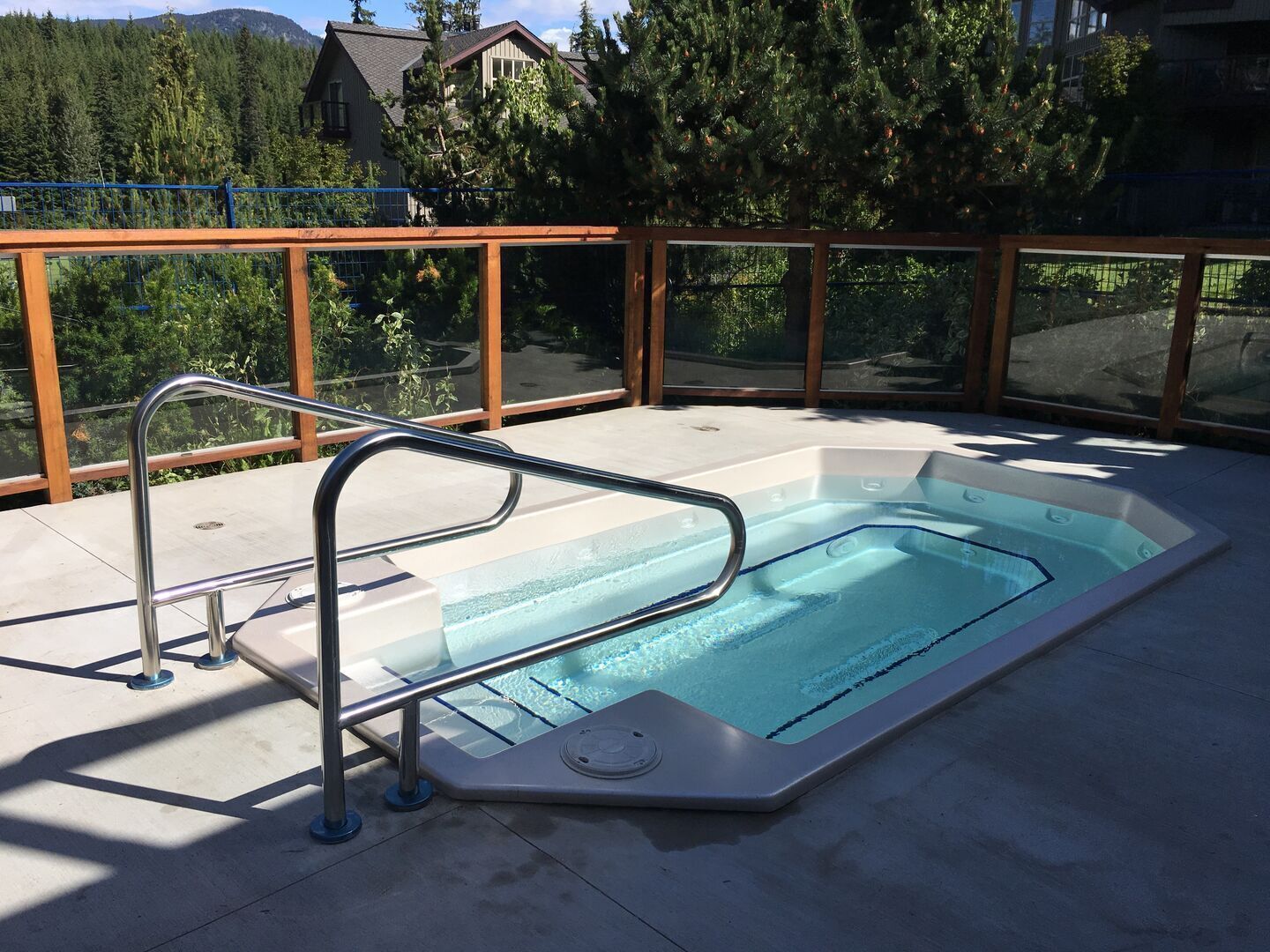 Un jacuzzi con barandilla metálica, rodeado por una cerca de madera y paneles de vidrio, con vistas a un área boscosa.