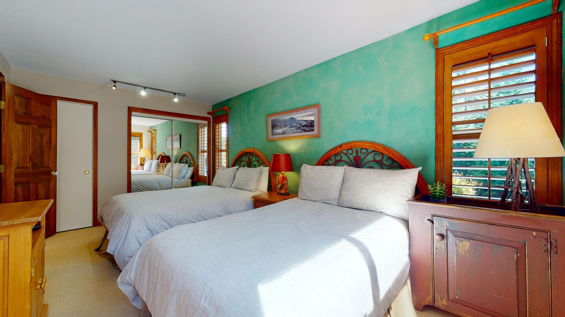 A room with two beds, a wooden wardrobe, a mirror, and a window with blinds.