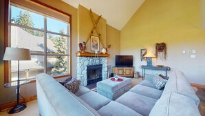 A living room with a stone fireplace, a sofa, a television, and a lamp. - 3BR | Blackcomb Greens | Wood Burning Fireplace | Complex Hot Tub | Parking | Mountain Views (Whistler)