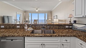 A modern kitchen with granite countertops, a double sink, and a dishwasher. There is a dining area with a table and chairs, and a large window offering a view of the ocean. - Boardwalk Beach Resort Family Getaway with Splash Zone and Gulf Views (Panama City Beach)