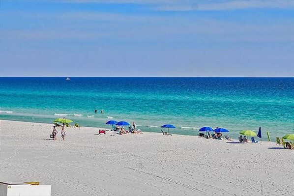 A beach with white sand, clear blue water, and several umbrellas providing shade.
