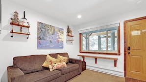 Ein gemütliches Wohnzimmer mit einem braunen Sofa, einem Gemälde einer verschneiten Landschaft und Holzregalen mit Weihnachtsdekorationen.