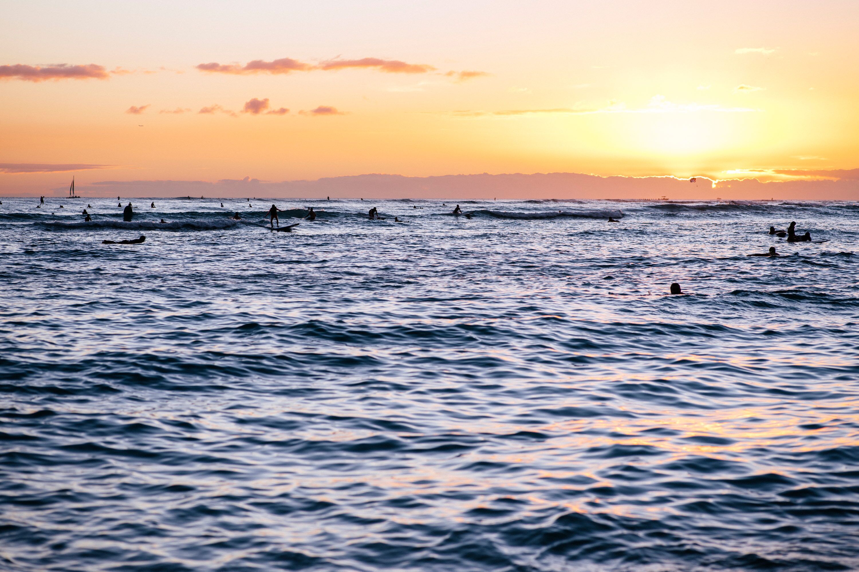 A sunset over a calm sea with several people swimming and a sailboat in the distance.