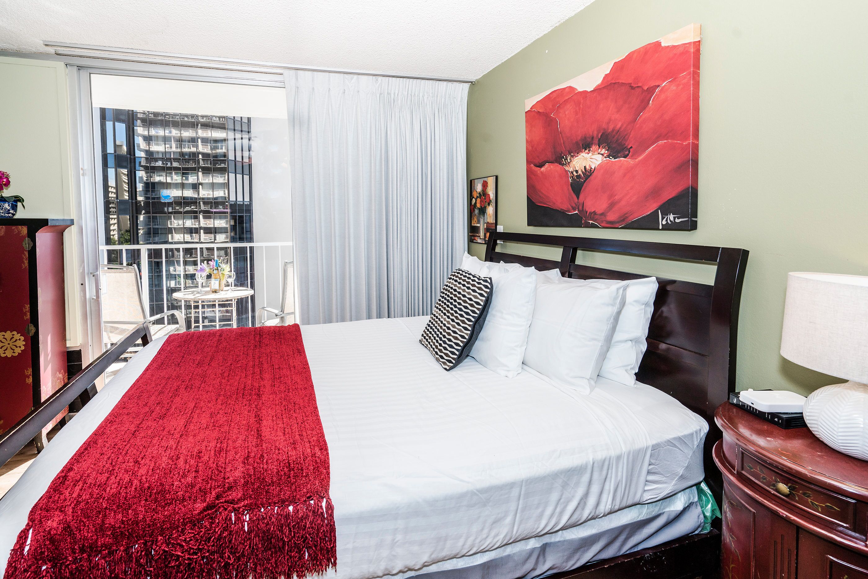 A hotel room with a large bed, a red throw blanket, white bedding, a red flower painting on the wall, and a view of a building through the window.