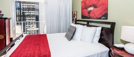 A hotel room with a large bed, a red throw blanket, white bedding, a red flower painting on the wall, and a view of a building through the window.