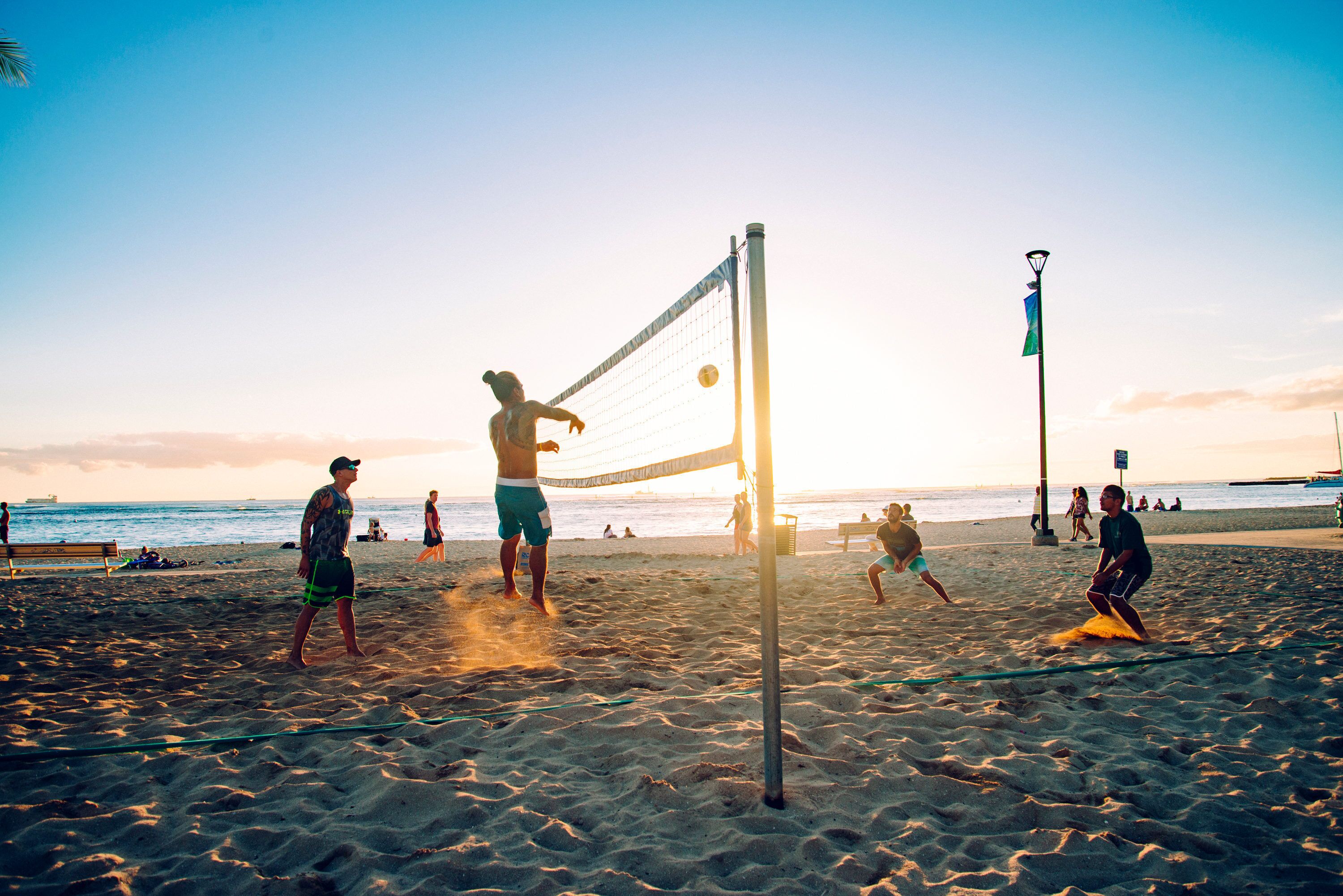 Ein Beachvolleyballspiel mit Spielern auf einem Sandplatz, einem Volleyballnetz und einem Sonnenuntergang im Hintergrund.