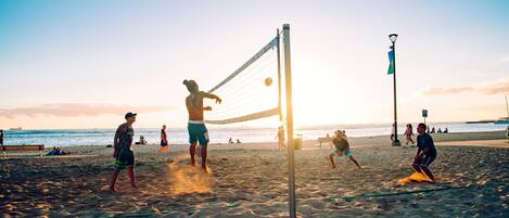 Ein Beachvolleyballspiel mit Spielern auf einem Sandplatz, einem Volleyballnetz und einem Sonnenuntergang im Hintergrund.