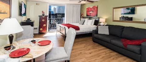 A hotel room with a bed, a sofa, a dining table with chairs, a balcony with a view of the city, and a ceiling fan.