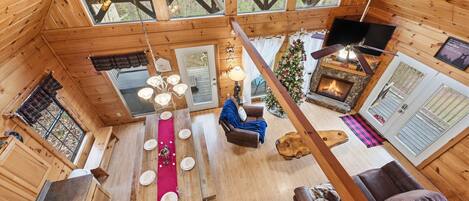 A cozy cabin interior with a dining area, a fireplace, and a large window overlooking the forest.