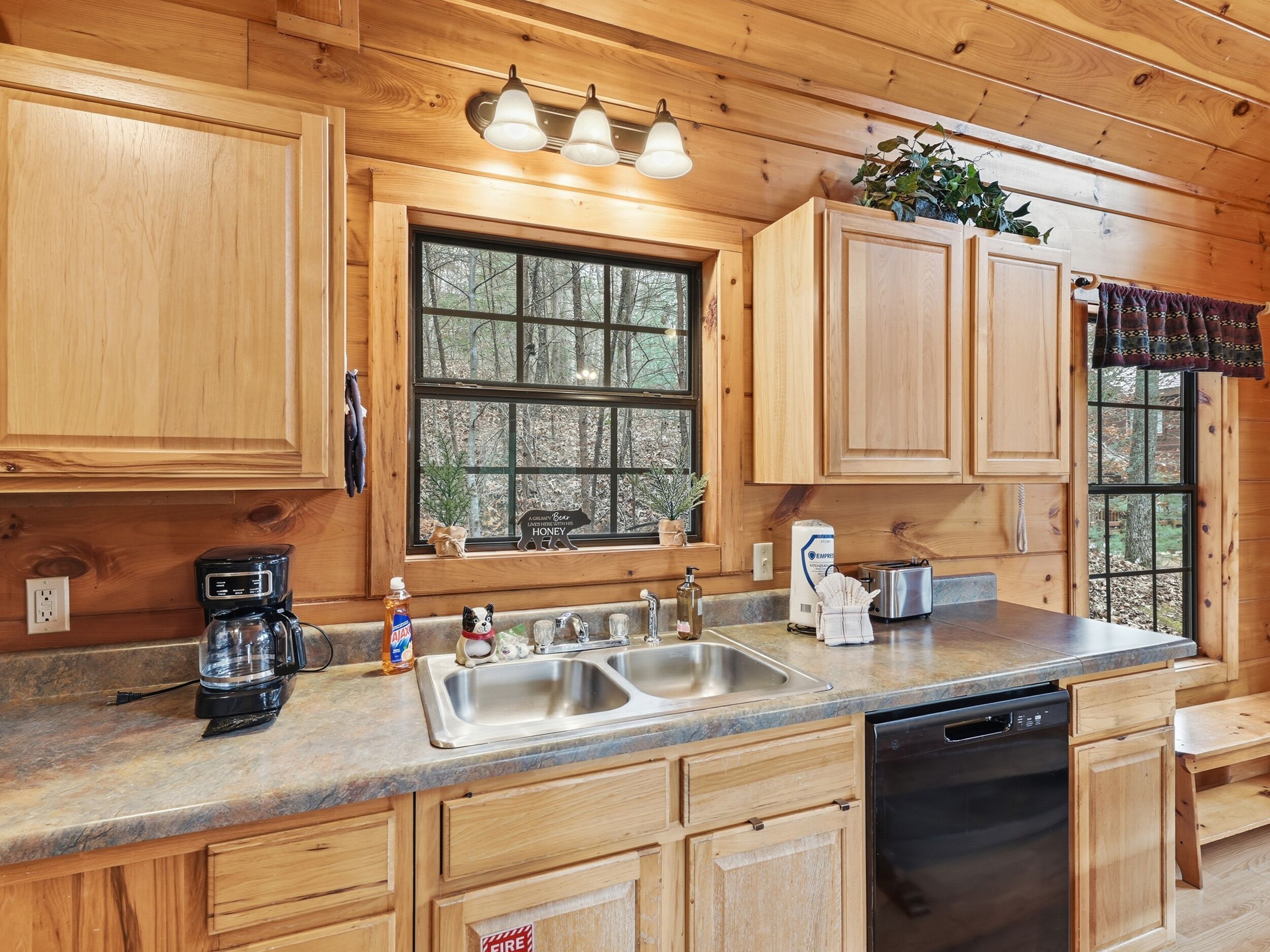 A wooden cabin kitchen with a large window, wooden cabinets, a sink, a coffee maker, and a dishwasher.