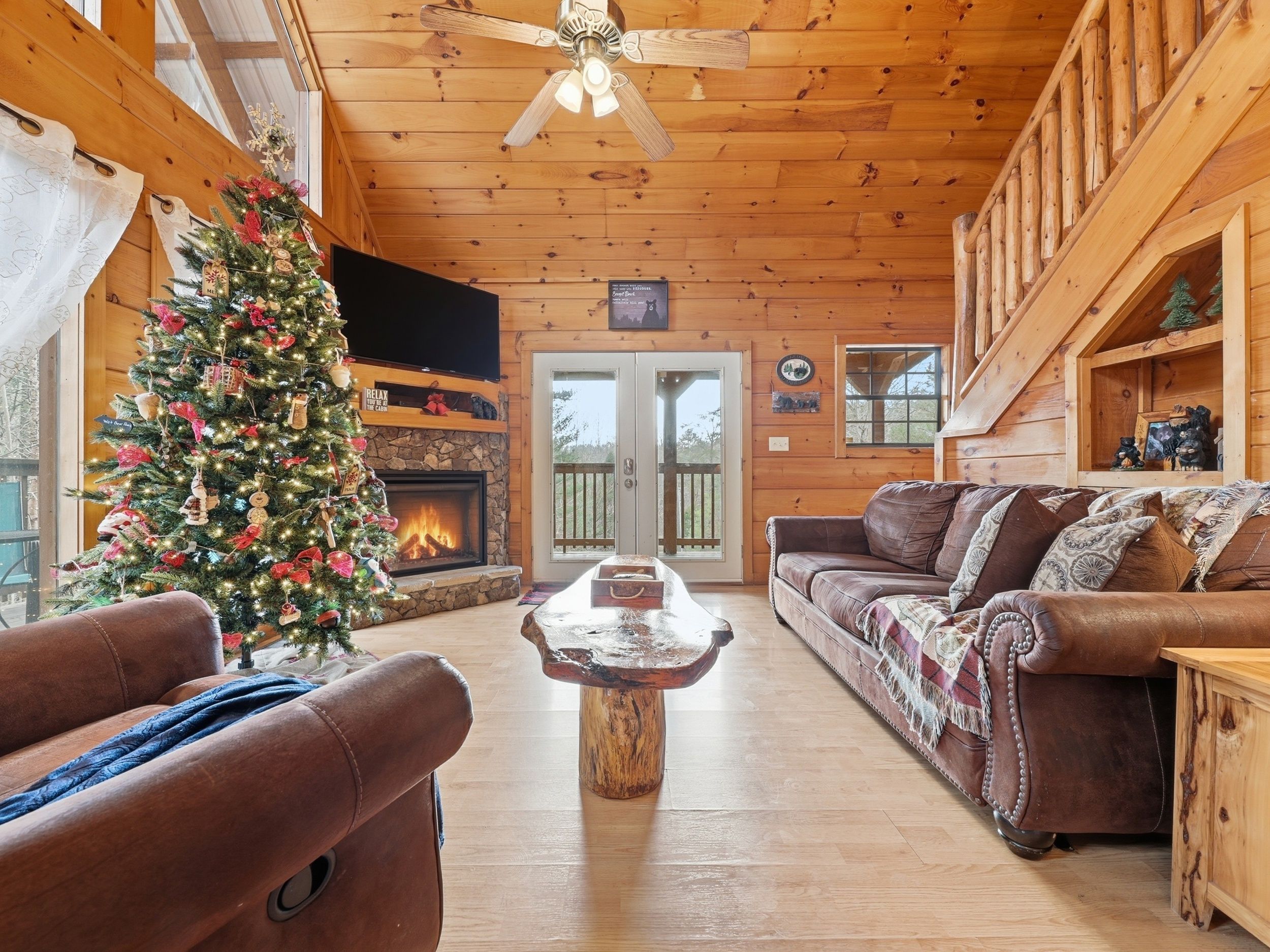 A cozy log cabin interior with a fireplace, a Christmas tree, leather sofas, and a stone fireplace surround.