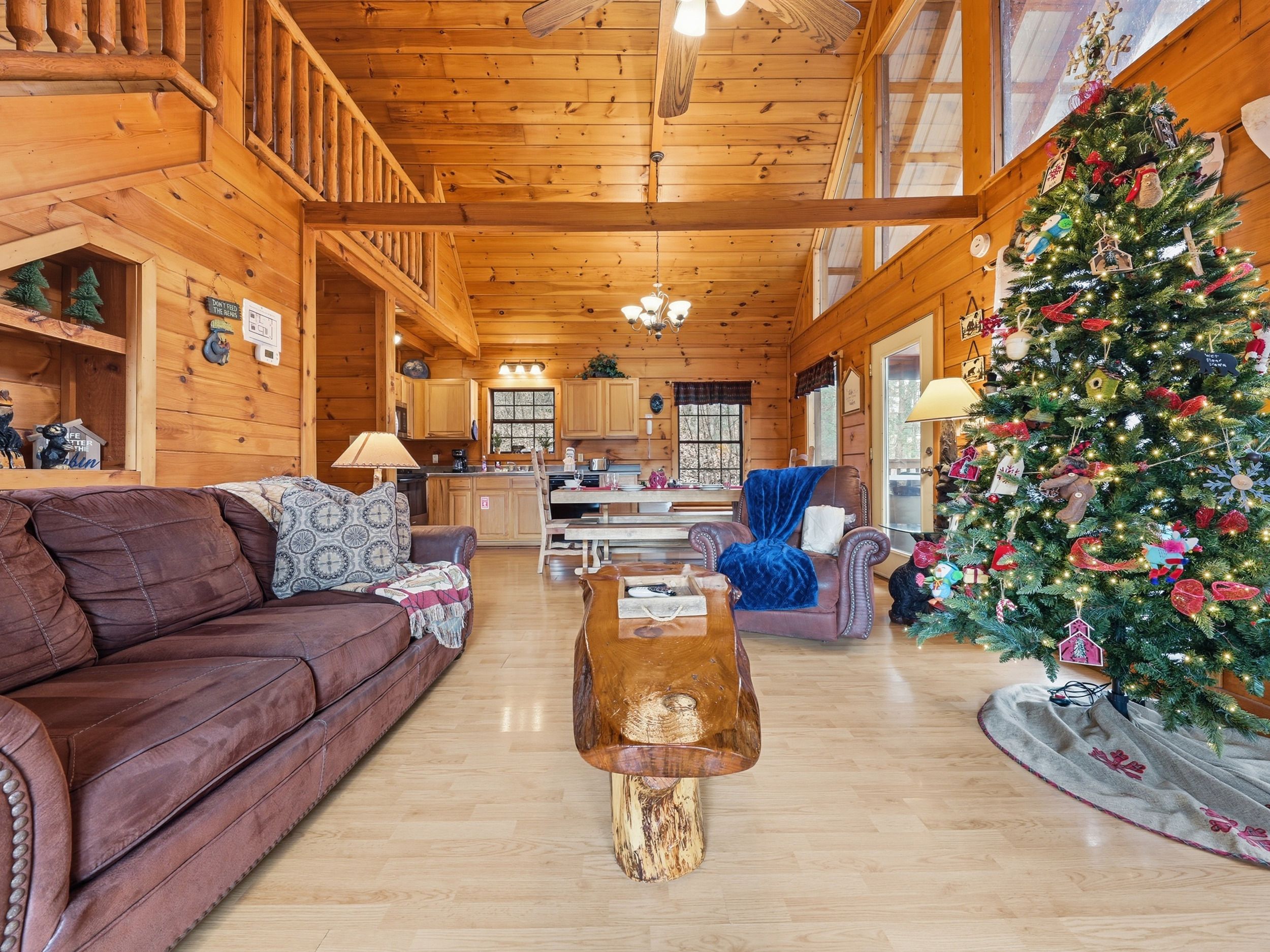 A cozy log cabin interior with a wooden staircase, a dining area, a living room with a leather sofa, and a decorated Christmas tree.