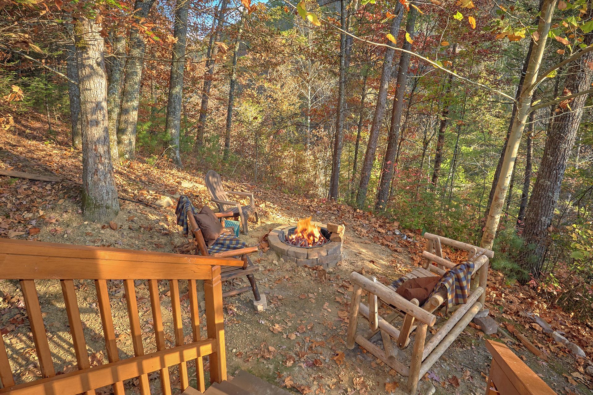Um camping arborizado com fogueira, bancos de madeira e um deck.