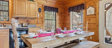 A wooden cabin interior with a dining table set for four, a kitchen area with wooden cabinets, and a large window providing a view of the outdoors.