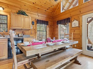 Ein Interieur einer Holzhütte mit einem Esstisch für vier Personen, einer Küchenzeile mit Holzausstattung und einem großen Fenster mit Blick ins Freie.