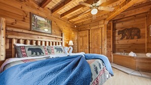 Une chambre à coucher dans un chalet en bois, avec un lit, un tableau, une tête de lit décorée avec un motif d’ours, une baignoire et un ventilateur de plafond.