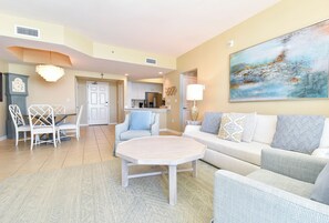 A spacious living room with a large painting, a dining area with white chairs, and a kitchen in the background. - Serene Beachfront Condo w/ Gulf Views & Close Dining in The Heart of Destin! 💙☀ (Destin)