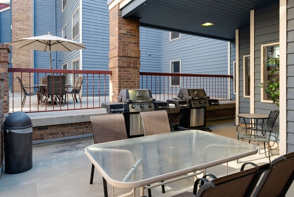 Un patio exterior con una mesa de vidrio, sillas de metal, una parrilla y un cubo de basura.