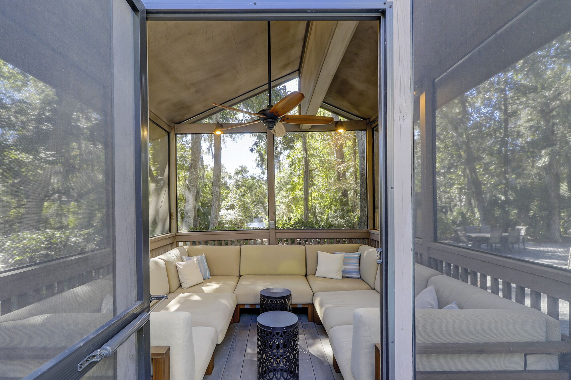 A screened porch with a ceiling fan, cushioned seating, and a view of trees.