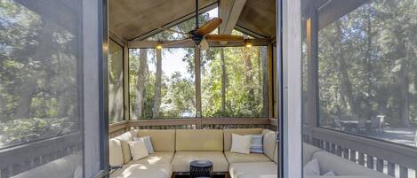 A screened porch with a ceiling fan, cushioned seating, and a view of trees.