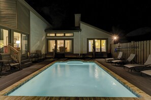 Ein Schwimmbecken mit Holzterrasse, Sitzgelegenheiten im Freien und ein Haus mit großen Fenstern.