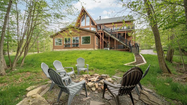 A log cabin with a staircase leading to a deck, surrounded by trees and a fire pit with chairs.