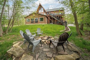 A log cabin with a staircase leading to a deck, surrounded by trees and a fire pit with chairs. - Cozy Rooms, Indoor Pool, Lake Perks & Hot Tub (McHenry)
