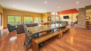 A spacious living room with a large wooden table, comfortable seating, a stone fireplace, and large windows.