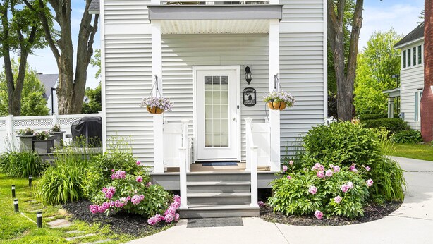 Et toetagers hus med en veranda, en postkasse og en lille have med lyserøde blomster.