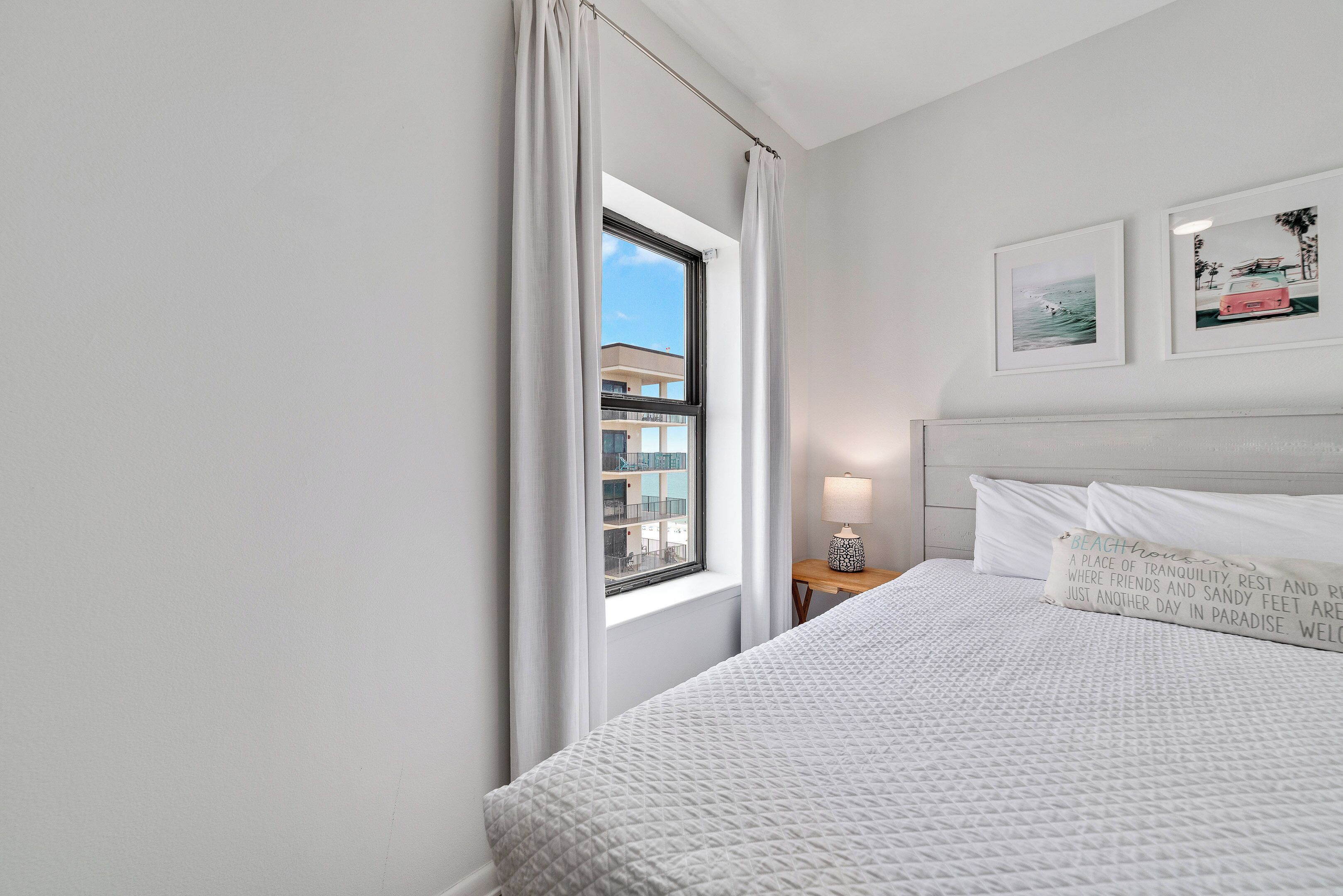 Une chambre d’hôtel avec un grand lit, une fenêtre avec des rideaux, une table de chevet avec une lampe et deux tableaux encadrés au mur.