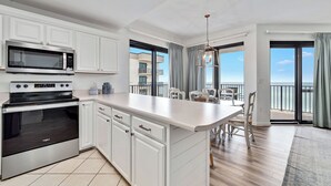 Une cuisine moderne avec des meubles blancs, un four à micro-ondes encastré et un four traditionnel. On y trouve un coin repas avec une table et des chaises, ainsi qu’un balcon offrant une vue sur la mer.