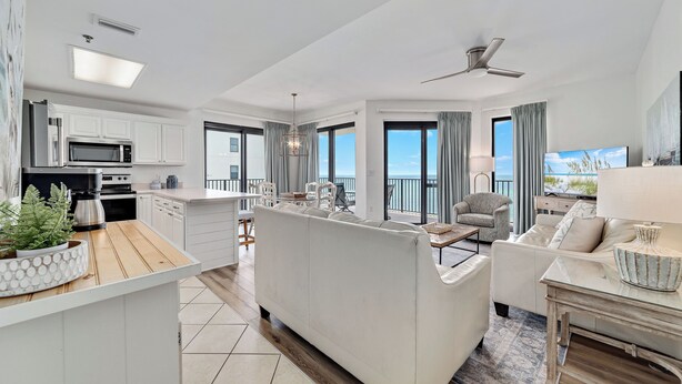 Un appartement moderne comprenant une cuisine, un coin repas et un salon. Il est doté d’un grand îlot central, de meubles blancs et de grandes fenêtres offrant une vue dégagée.