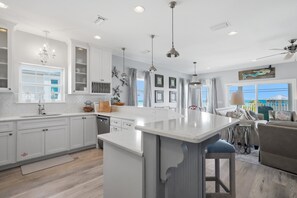 Une cuisine spacieuse avec des meubles blancs, un îlot central, des luminaires suspendus et une vue sur l’extérieur.