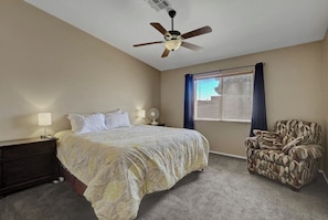 Une chambre à coucher comprenant un lit, un ventilateur de plafond, une table de chevet, une chaise et une fenêtre avec des stores.