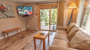 Un salon comprenant un canapé, une table basse en bois, un banc, un téléviseur fixé au mur et une porte-fenêtre coulissante donnant sur un espace extérieur.