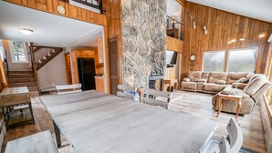 Un vaste espace de vie comprenant une grande table en bois, un mur décoratif en pierre et un coin salon confortable avec une cheminée.