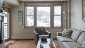 A cozy living room with a fireplace, a sofa, a coffee table with chess pieces, and a view of a snowy landscape through large windows. - Zephyr Mountain Lodge 2315 | Ski In Ski Out | Walk To Base Area (Winter Park)