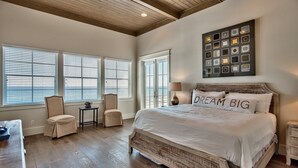 A spacious bedroom with a large bed, two chairs, a small table, and a view of the sea through large windows.