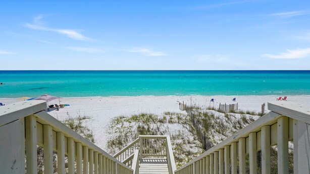A beach view from a wooden staircase leading to a sandy beach with clear turquoise water.