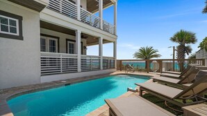 A pool area with a clear view of the ocean, a covered seating area, and a two-story building with balconies.