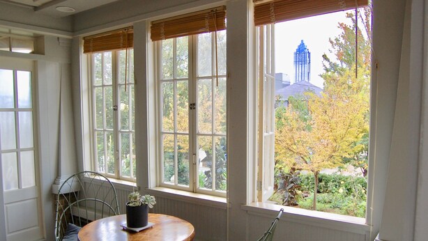 Una habitación con una mesa redonda de madera, dos sillas de metal y una planta en maceta. Amplias ventanas con marcos blancos y persianas de madera dejan ver árboles y un edificio al exterior.
