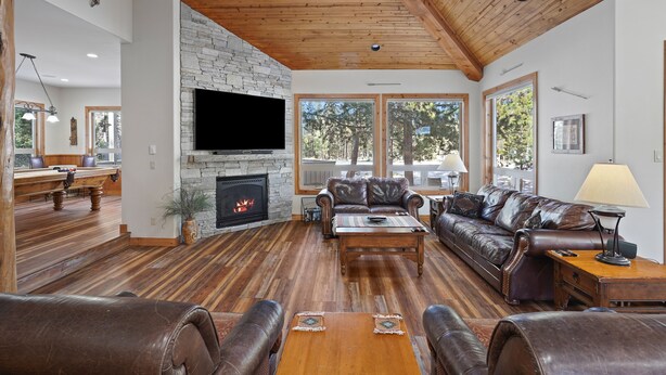 A spacious living room with a stone fireplace, leather sofas, wooden flooring, and large windows offering a view of the outdoors.